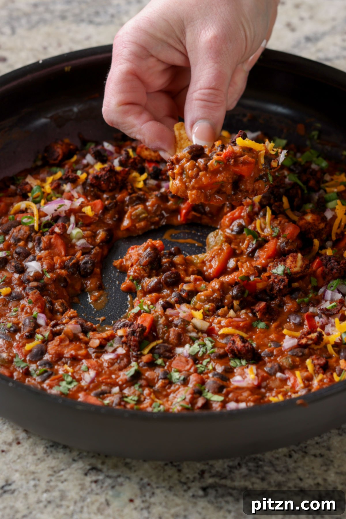 A hand dipping a crispy tortilla chip into a steaming pan of black beans and red lentils, highlighting the rich texture and inviting warmth of the dish.