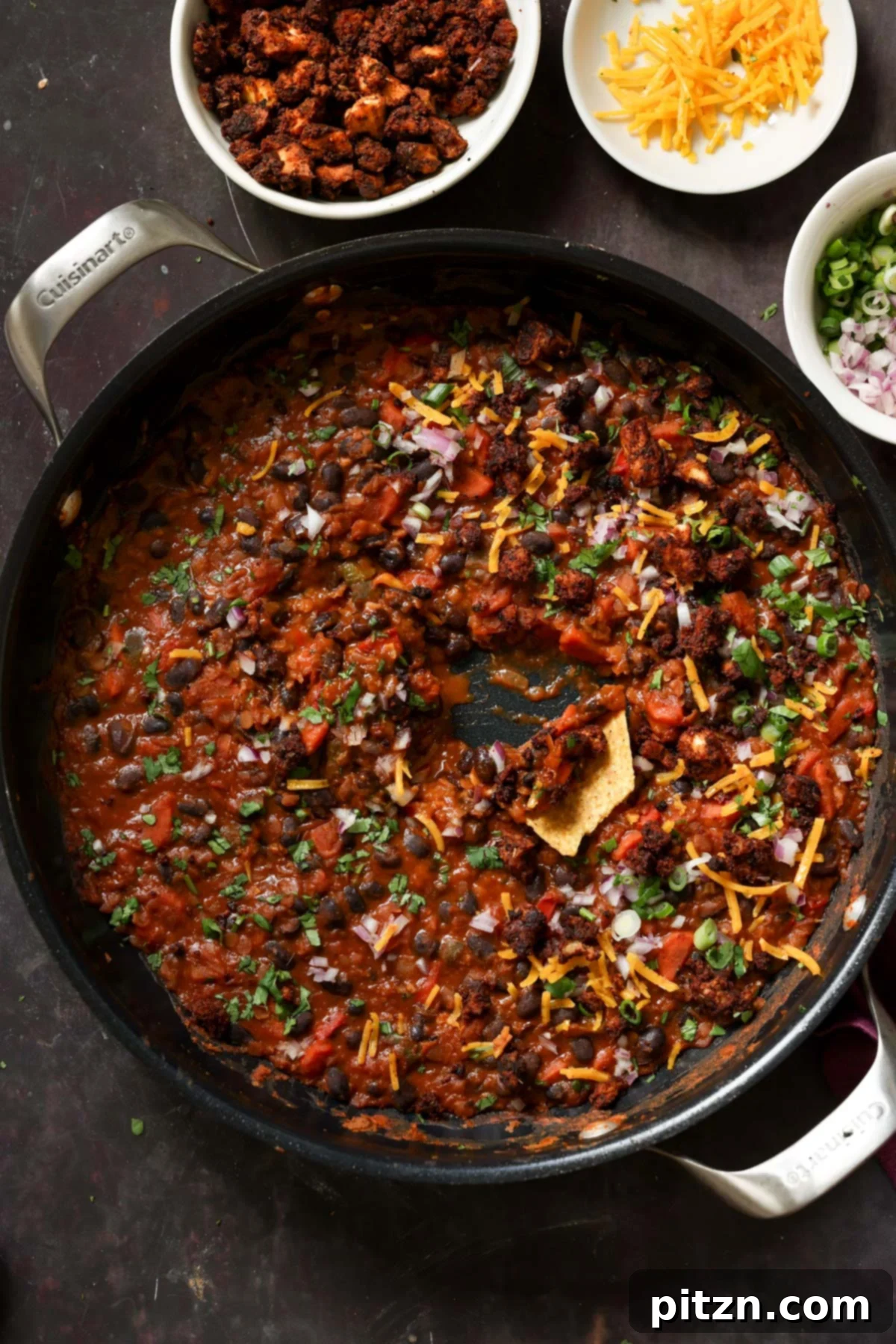 A close-up view of the rich black bean and lentil stew in a pan, now fully topped with fresh garnishes like cilantro, with a crispy tortilla chip being dipped in.