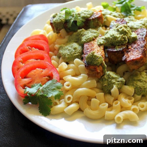 A bowl of pasta with spicy blackened tempeh strips, topped with fresh cilantro almond pesto and sliced tomatoes. The dish is vibrant and appetizing.