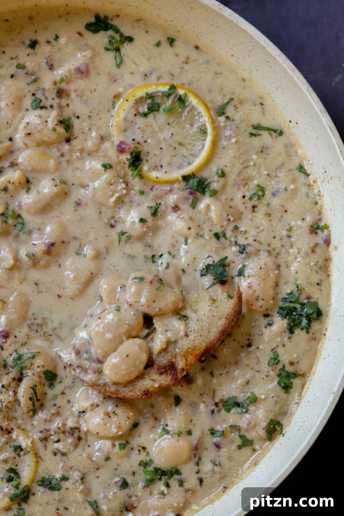 Close-up of a hand dipping crusty sourdough bread into a rustic pan of creamy lemon garlic butter beans, highlighting the rich sauce and tender beans.