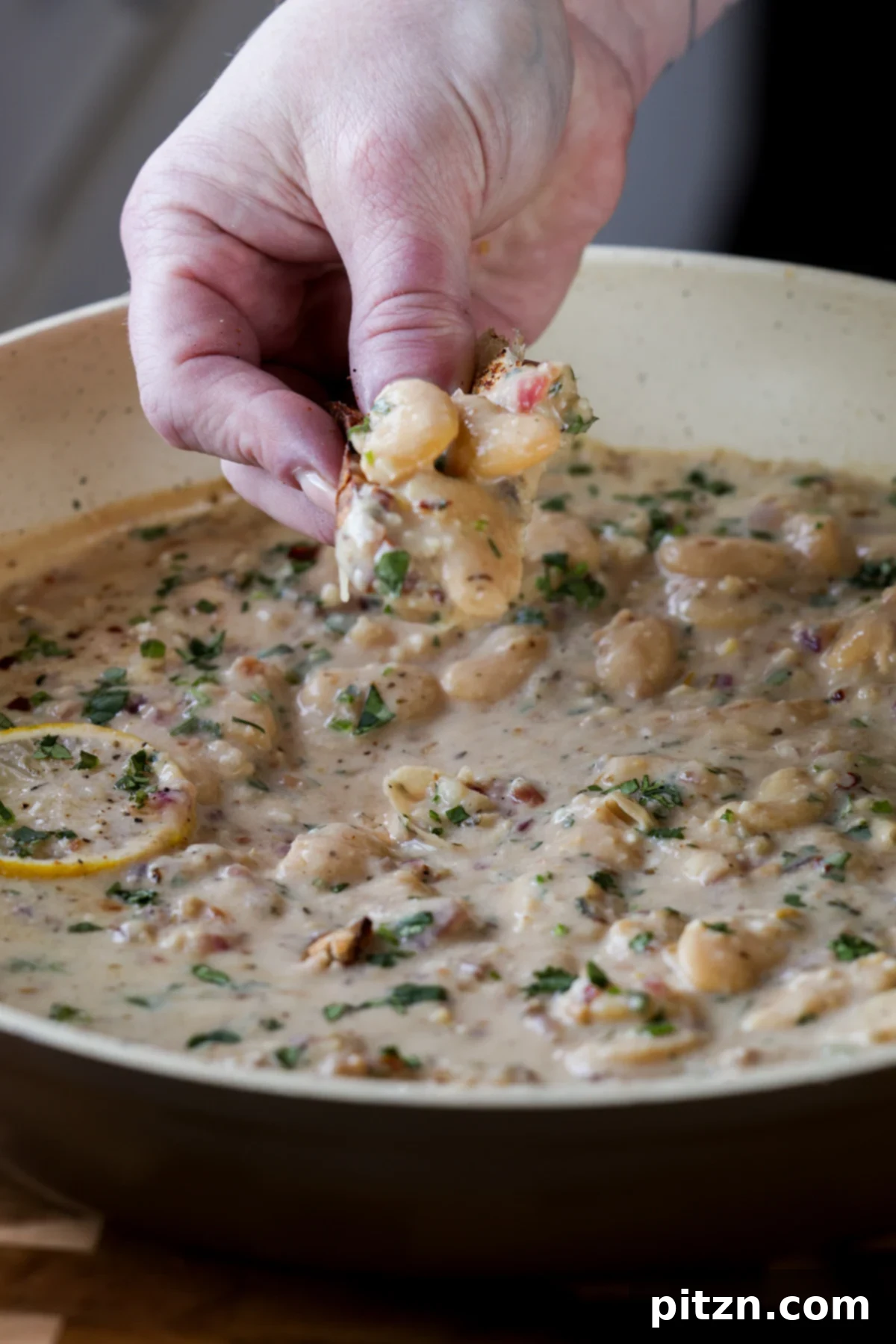 A hand scooping up creamy lemon garlic butter beans onto a piece of toasted bread, emphasizing the rich texture and delicious pairing.