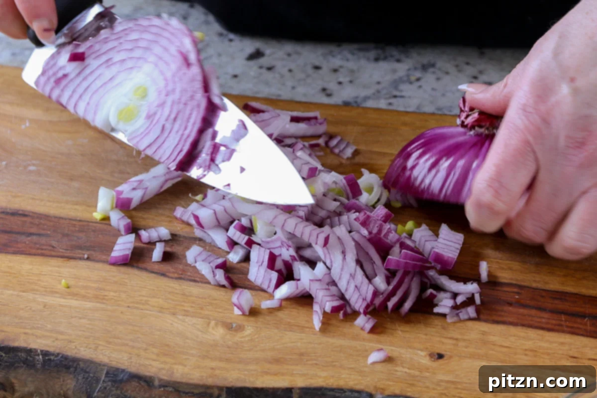 A sharp chef's knife finely chopping a white onion on a wooden cutting board, preparing the aromatics for the sauce.