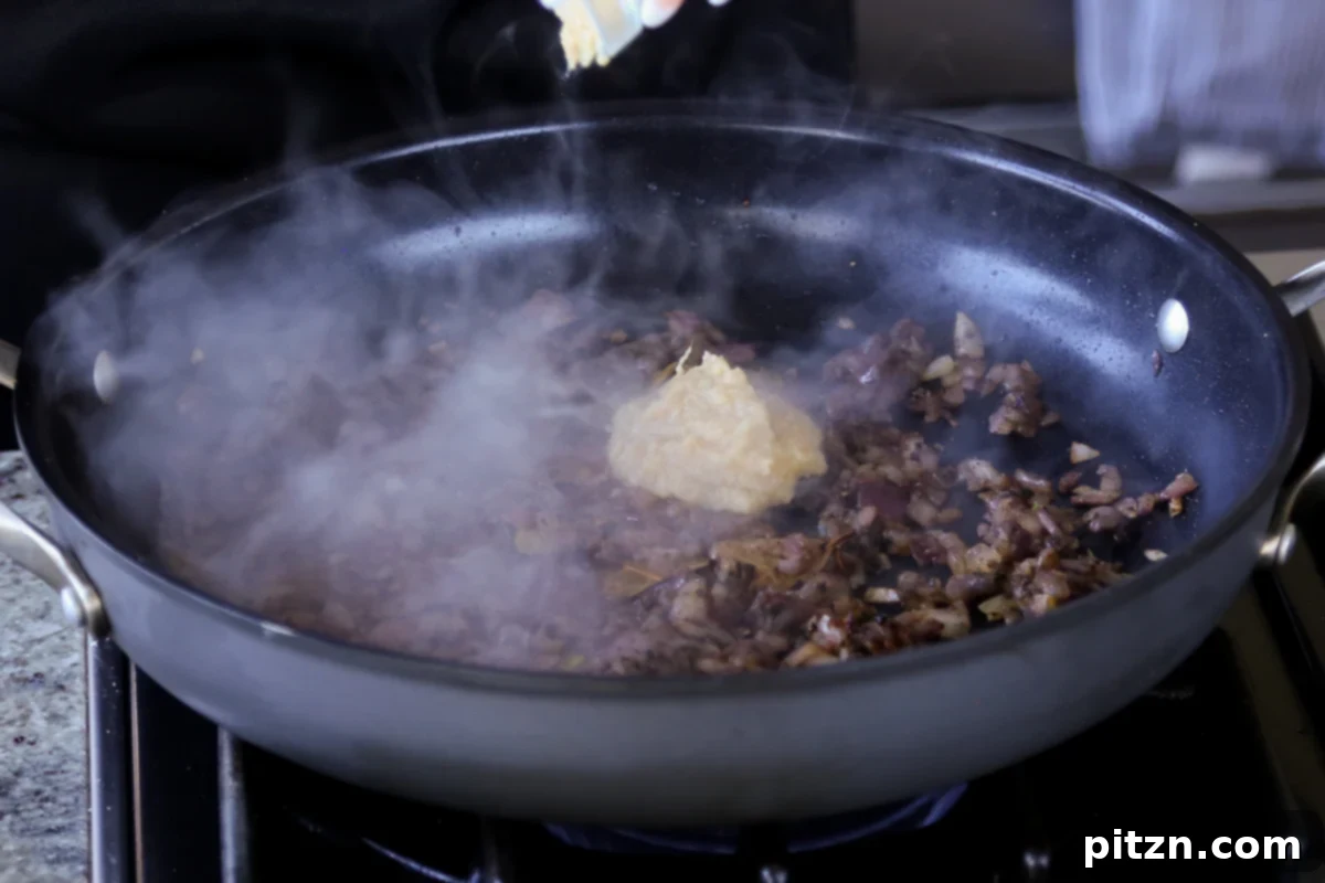 Ginger garlic paste being added to the cooked onion and spice mixture in the skillet, ready to be incorporated.