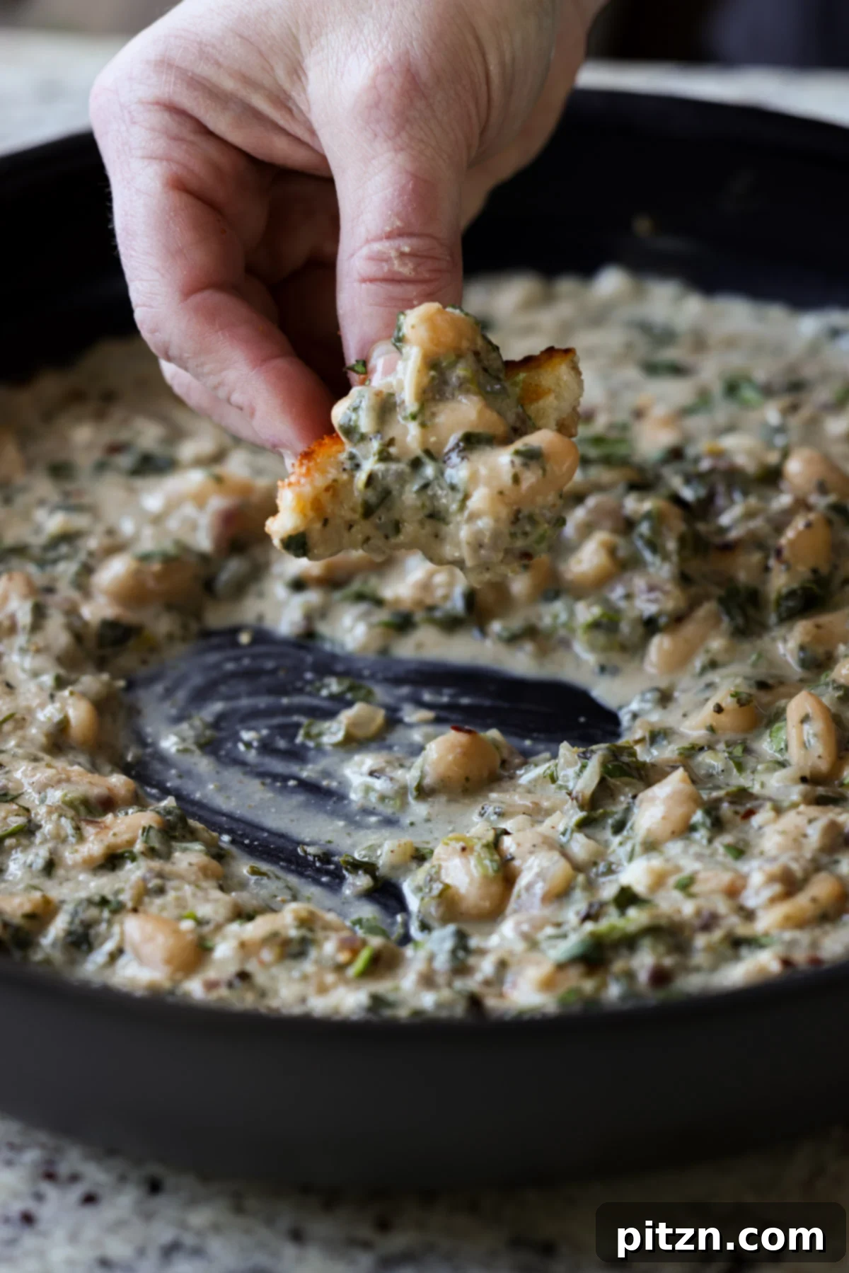 A hand scooping up a portion of creamy Kali Mirch Beans with a piece of toasted sourdough bread, showcasing the rich texture of the sauce and the tender beans.