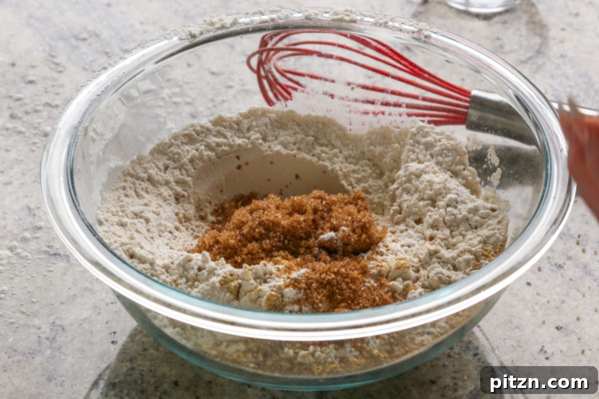 Luscious Triple Ginger Vegan Gingerbread Cake 10 A hand adding brown sugar from a small bowl into a larger bowl containing other dry ingredients for vegan gingerbread cake, ensuring all components are ready for mixing.