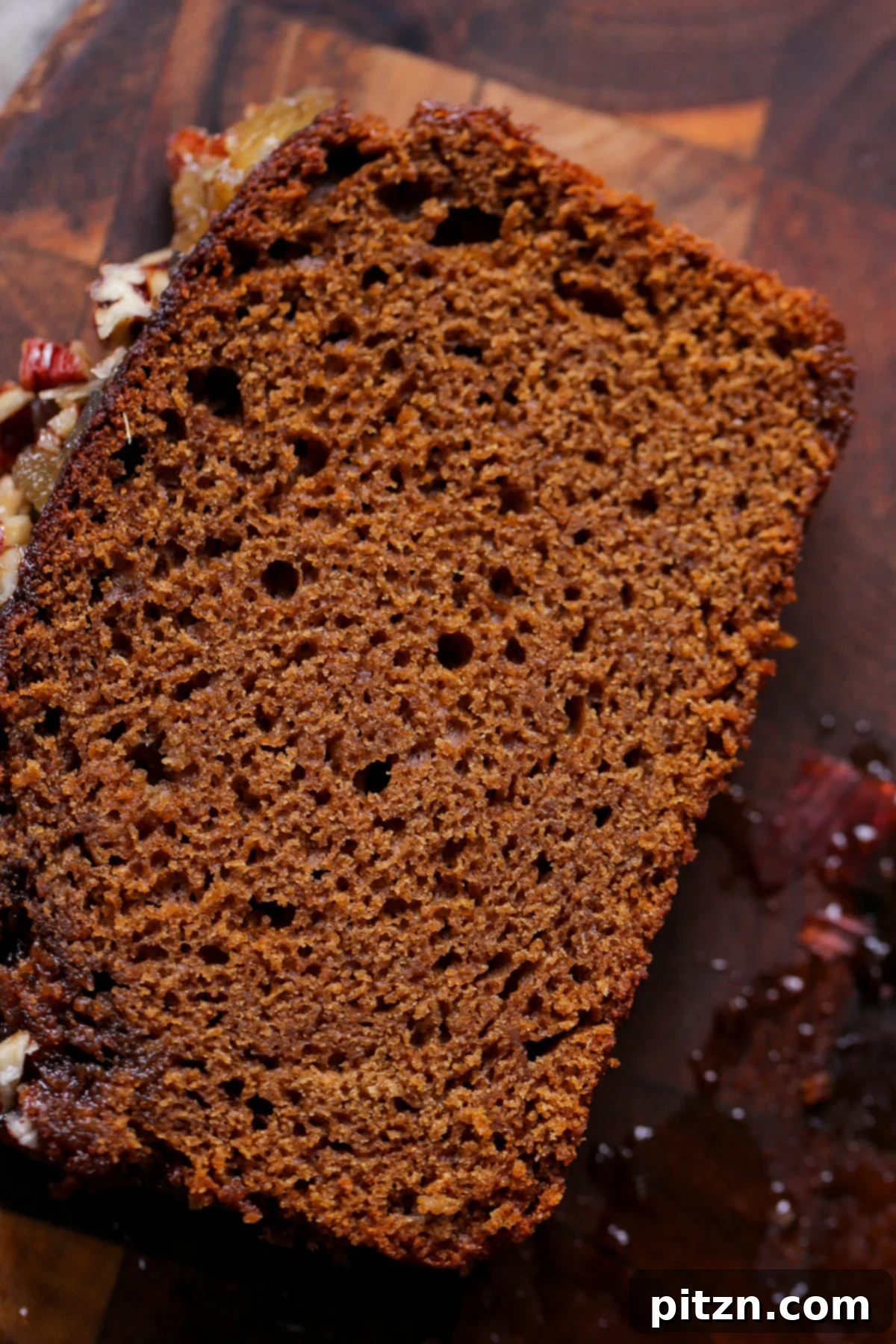 Luscious Triple Ginger Vegan Gingerbread Cake 6 A close-up of a thick slice of vegan gingerbread cake, artfully placed on a wooden cutting board. The top is generously coated with ginger glaze and studded with chopped nuts and candied ginger, highlighting its inviting texture.