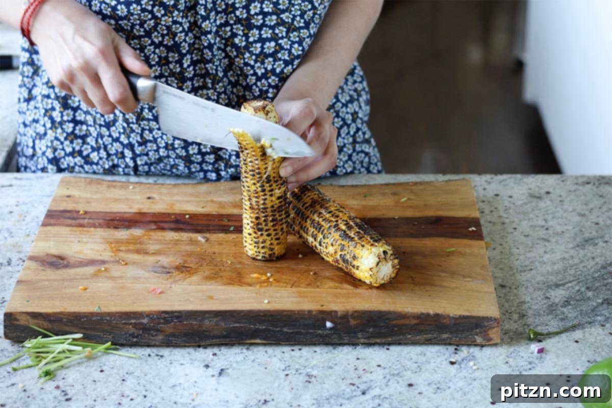 Tropical Chickpea & Mango Salad with Spiced Lime-Pepper Drizzle 9 A hand using a knife to slice roasted corn kernels off the cob into a bowl, with the charred cob visible.