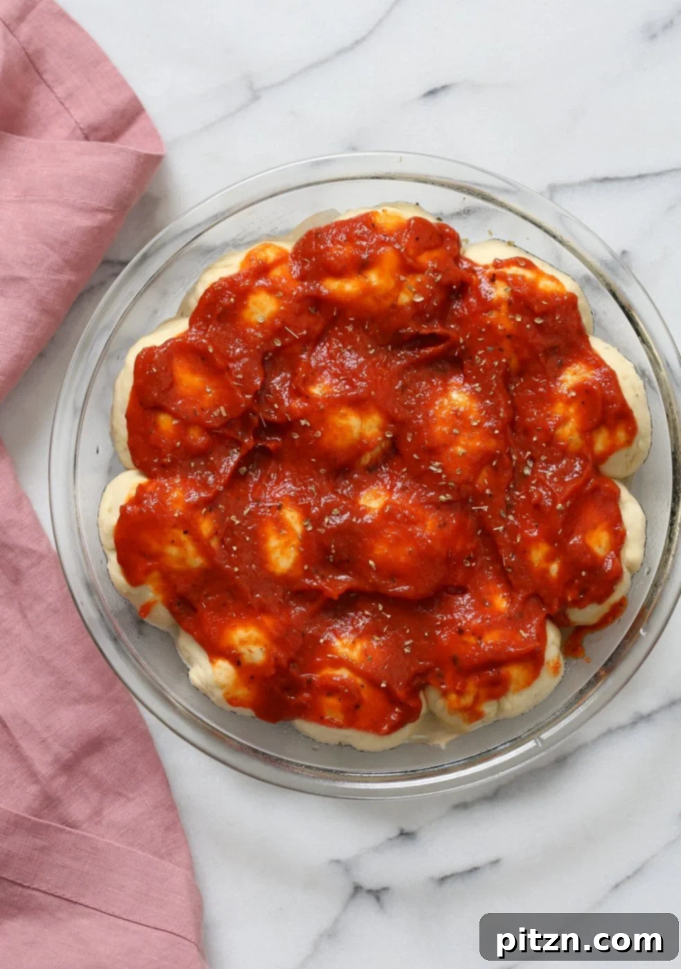 Cheesy Pizza Monkey Bread 7 Close-up of pizza dough balls in a pan with cashew cream poured between them, topped with pizza sauce, oregano, and pepper, prepped for baking Cheesy Pull Apart Pizza Bread.