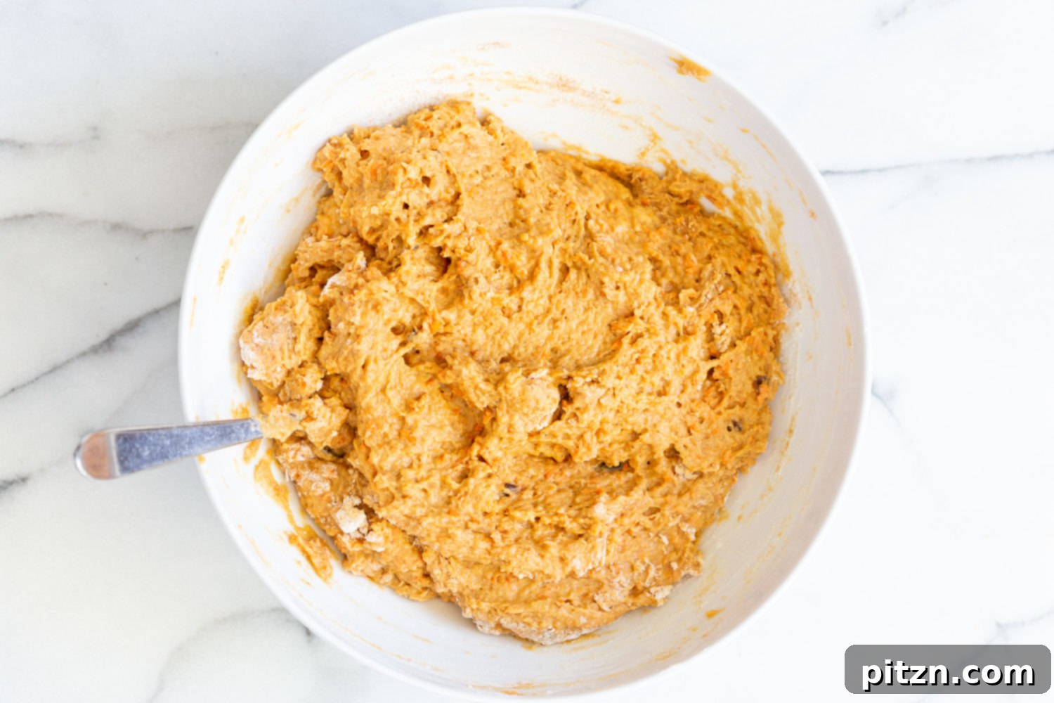 Stiff vegan carrot bread batter in a mixing bowl with a spoon, demonstrating the desired thick consistency.