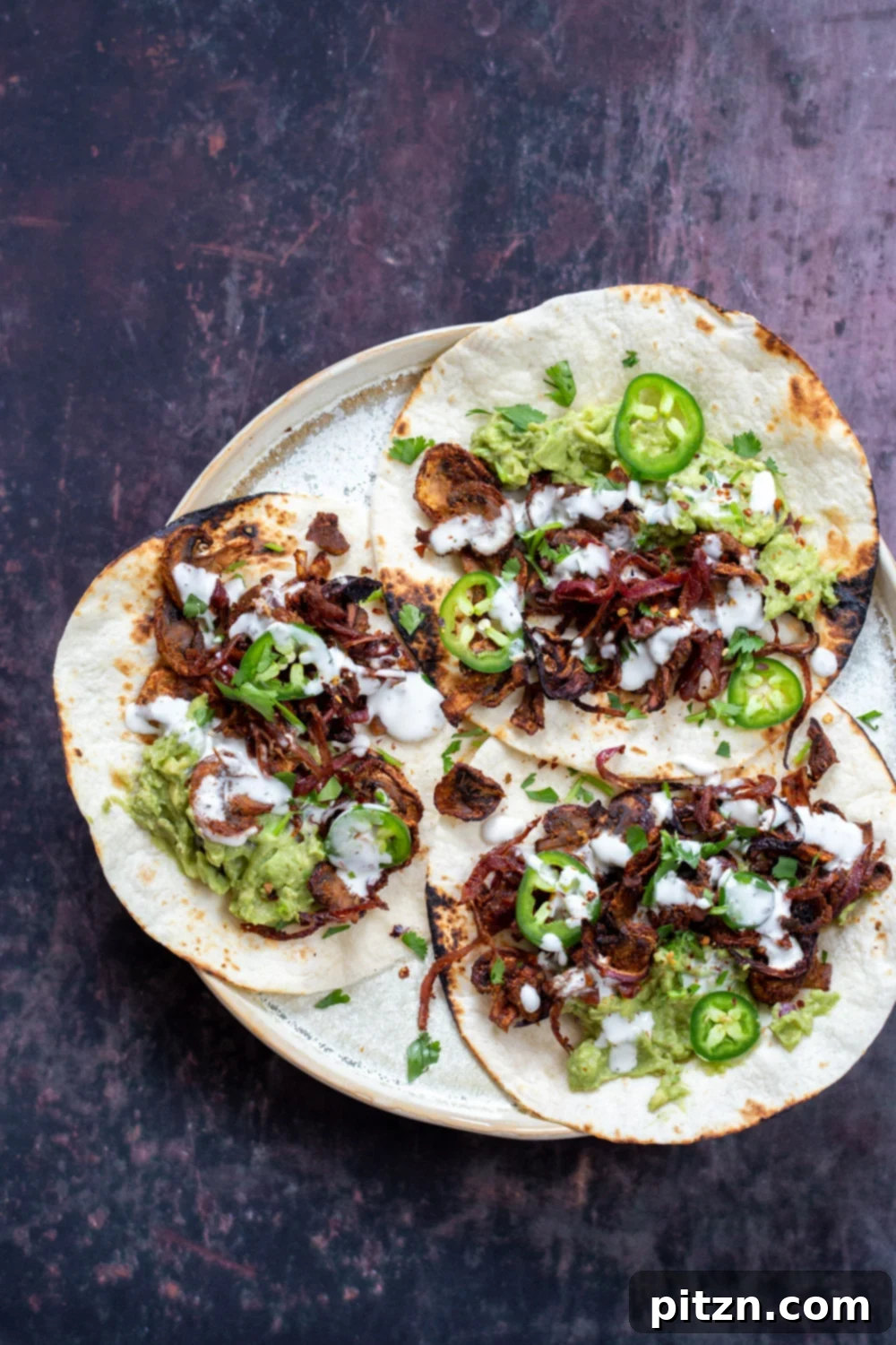 Three delicious Vegan Carnitas tacos filled with smoky mushroom mixture, topped with creamy guacamole, fresh jalapenos, and a dollop of vegan sour cream, artfully arranged on a speckled plate.