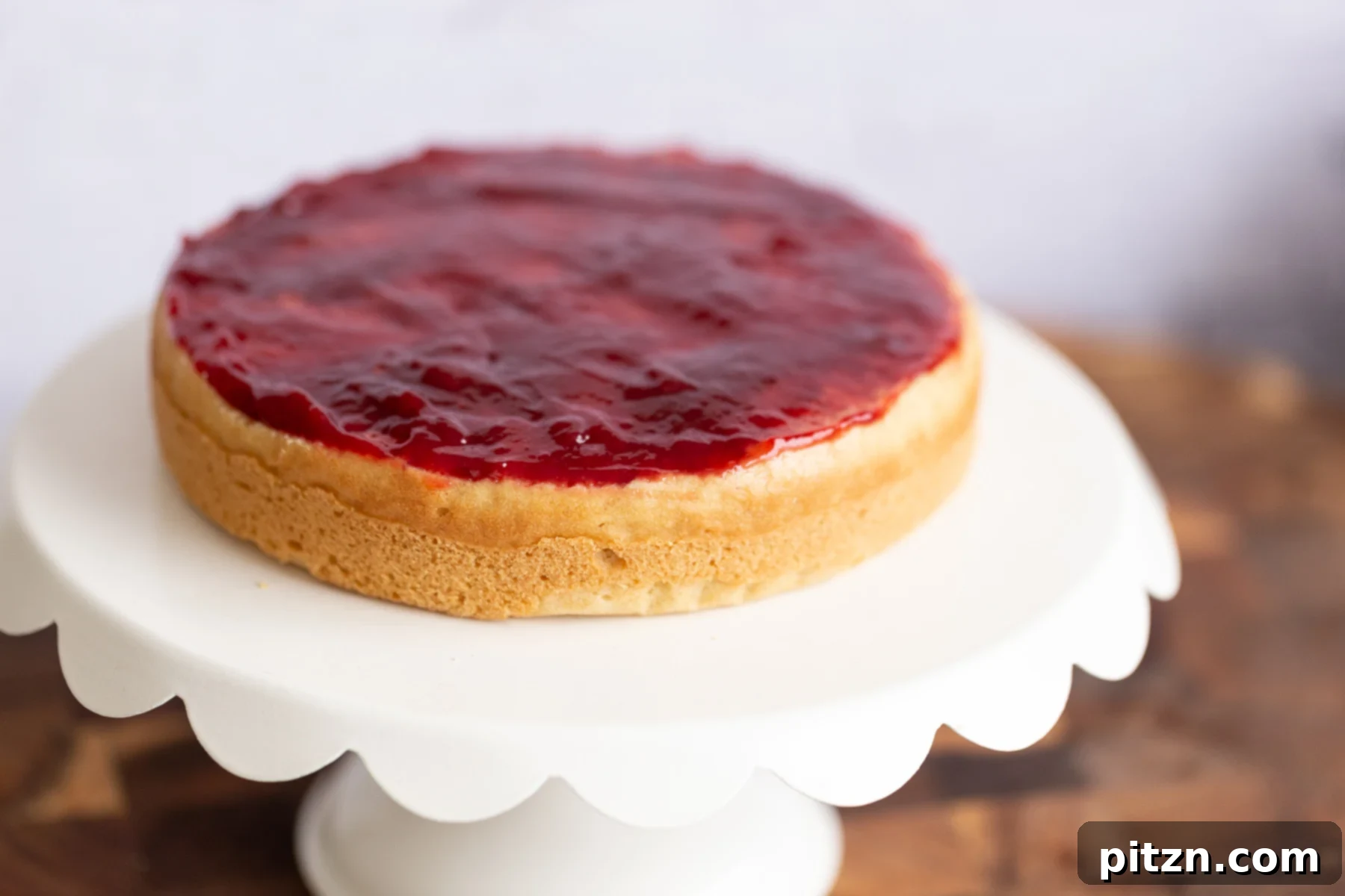 A delicate vegan sponge cake layer generously topped with smooth, vibrant raspberry jam, ready for the next layer.