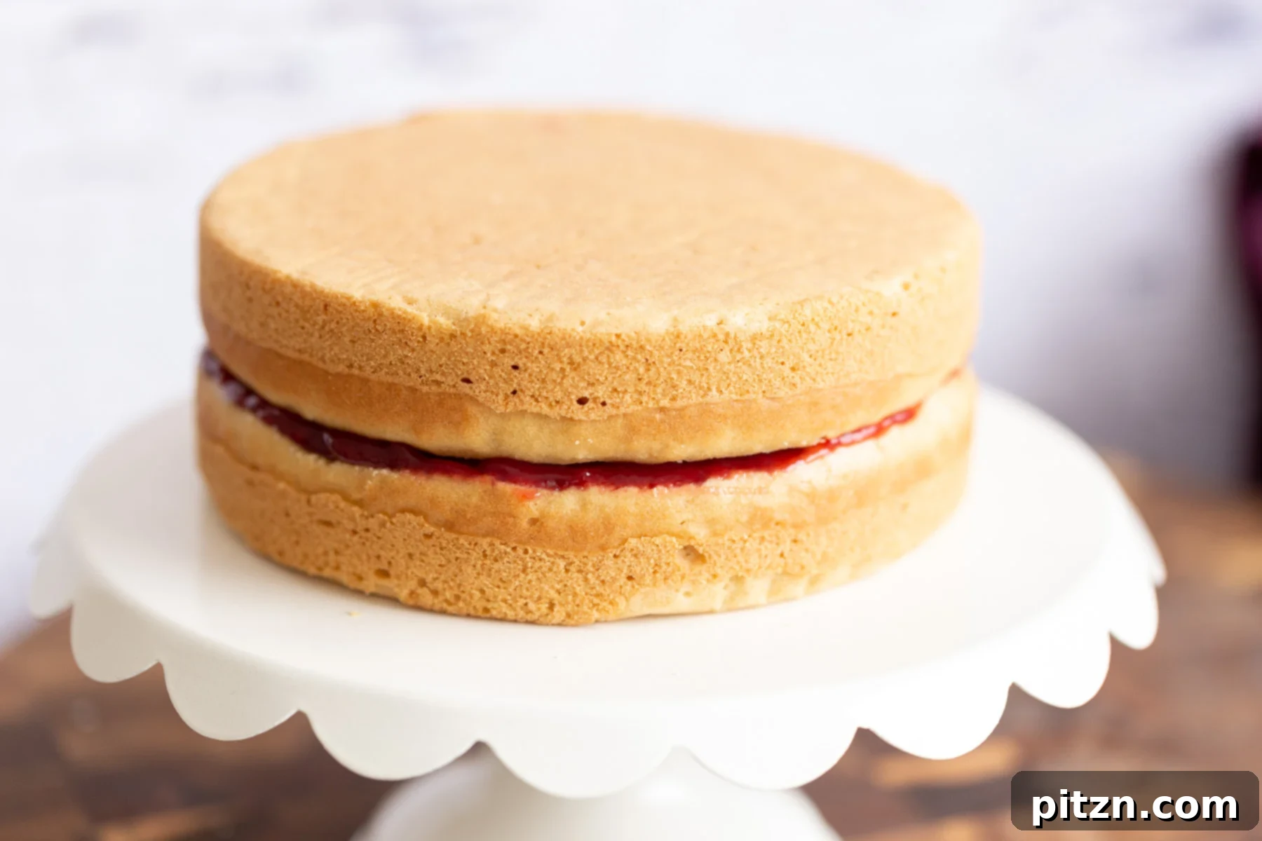 A fully assembled vegan white cake with two layers, filled with raspberry jam and topped with a creamy white frosting, on a cake stand.