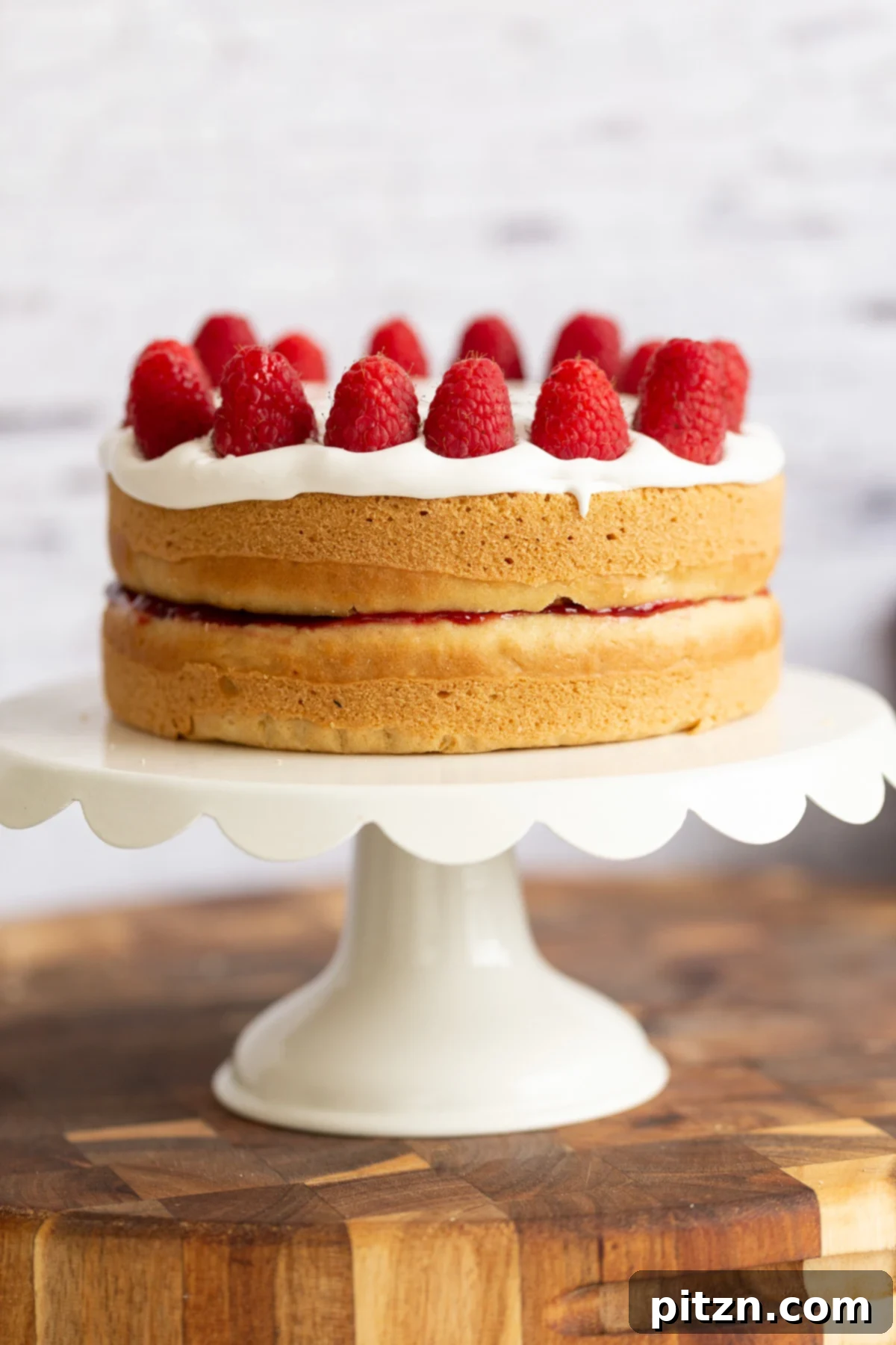 A stunning whole vegan white cake adorned with coconut whipped cream and fresh raspberries, presented on a pristine white cake stand.