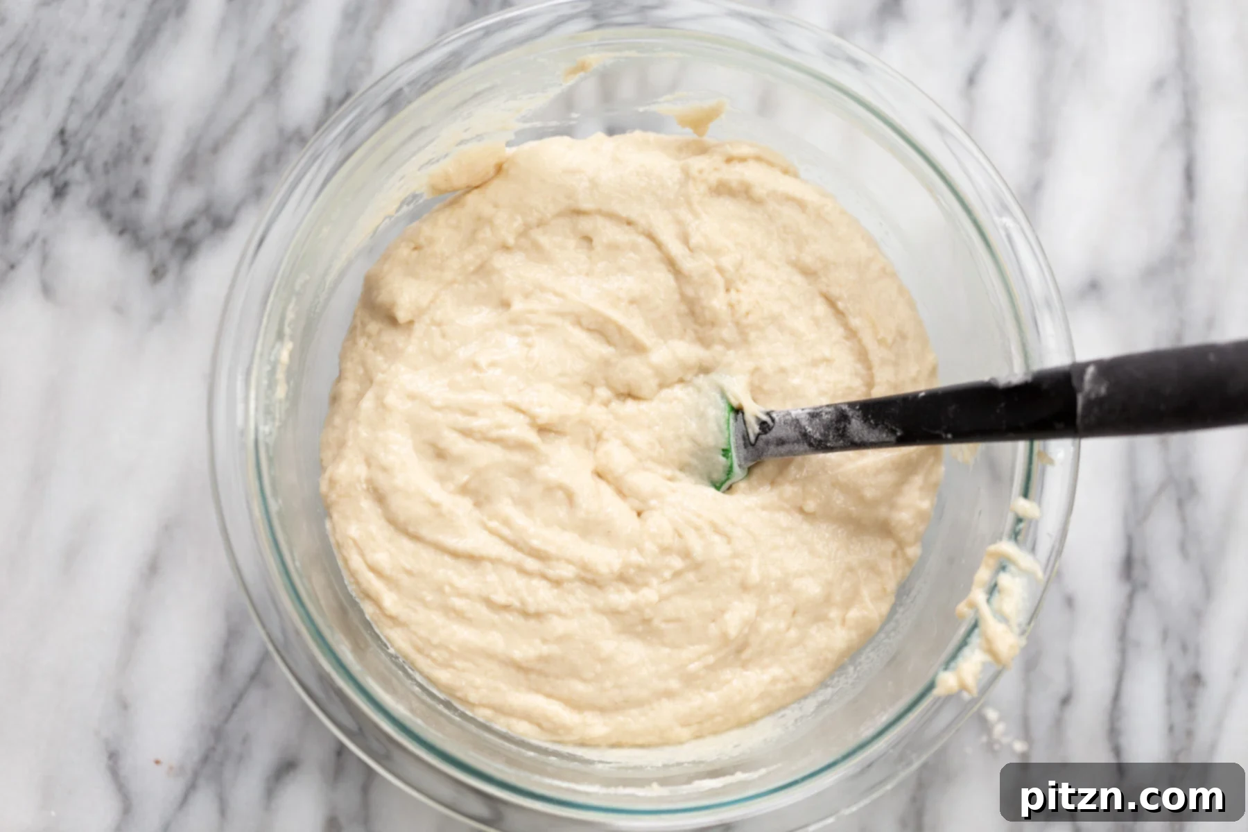 Vegan white cake batter being thoroughly mixed in a clear glass bowl, showcasing its smooth consistency.