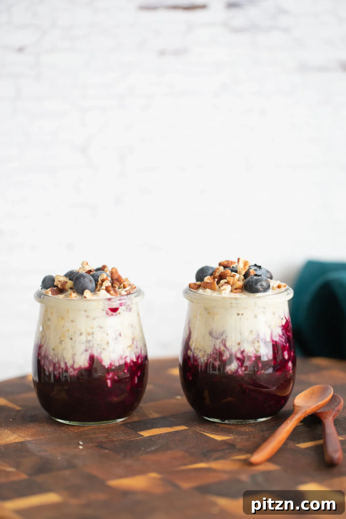 Easy Vegan Blueberry Overnight Oats Side view of two glass jars filled with layered blueberry overnight oats, topped with fresh blueberries and a dusting of lemon zest, resting on a wooden cutting board