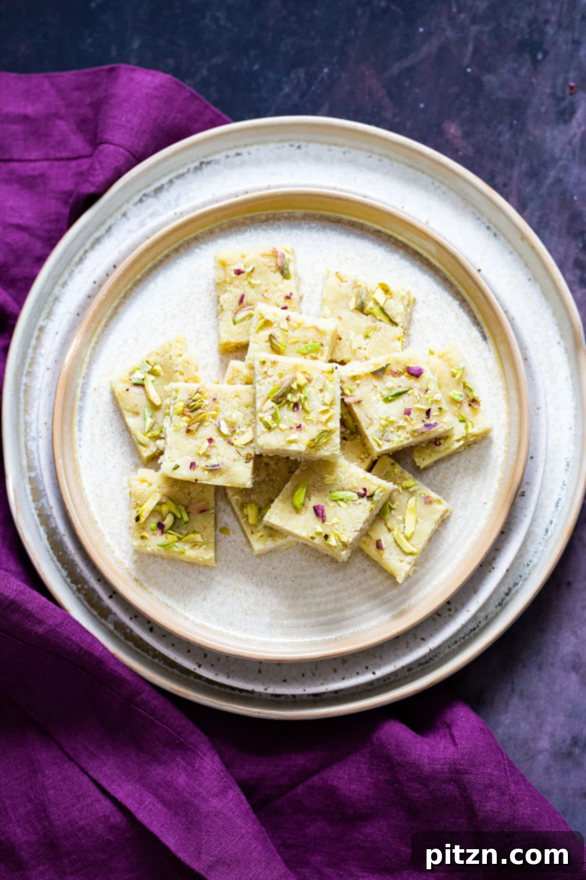 Vegan Almond Burfi with Pistachios A vibrant plate filled with freshly cut vegan almond burfi squares, elegantly garnished with chopped pistachios and delicate almond slivers. The texture appears smooth and fudgy.