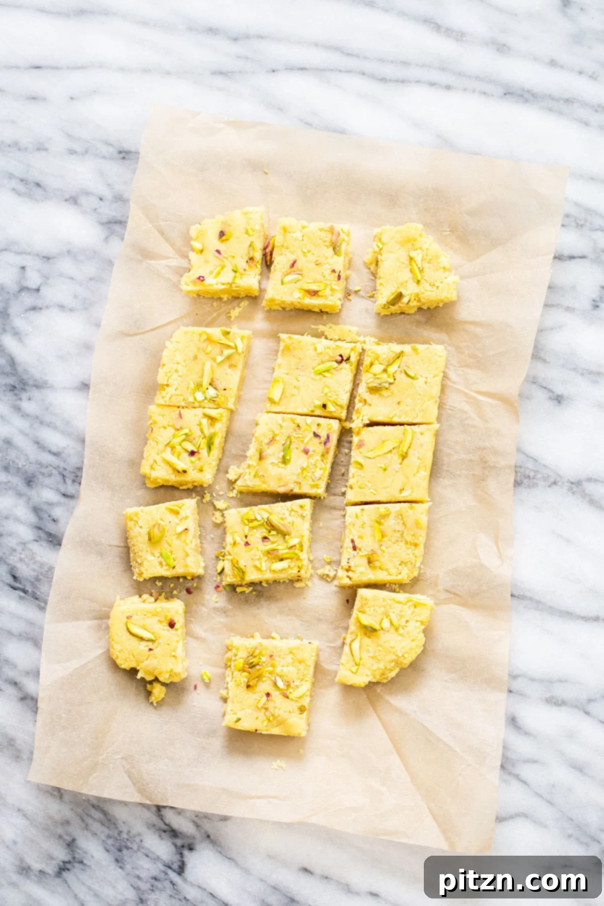 Homemade Vegan Almond Burfi A perfectly arranged row of vegan almond burfi squares sits on parchment paper, showcasing their smooth, light brown color and inviting texture.