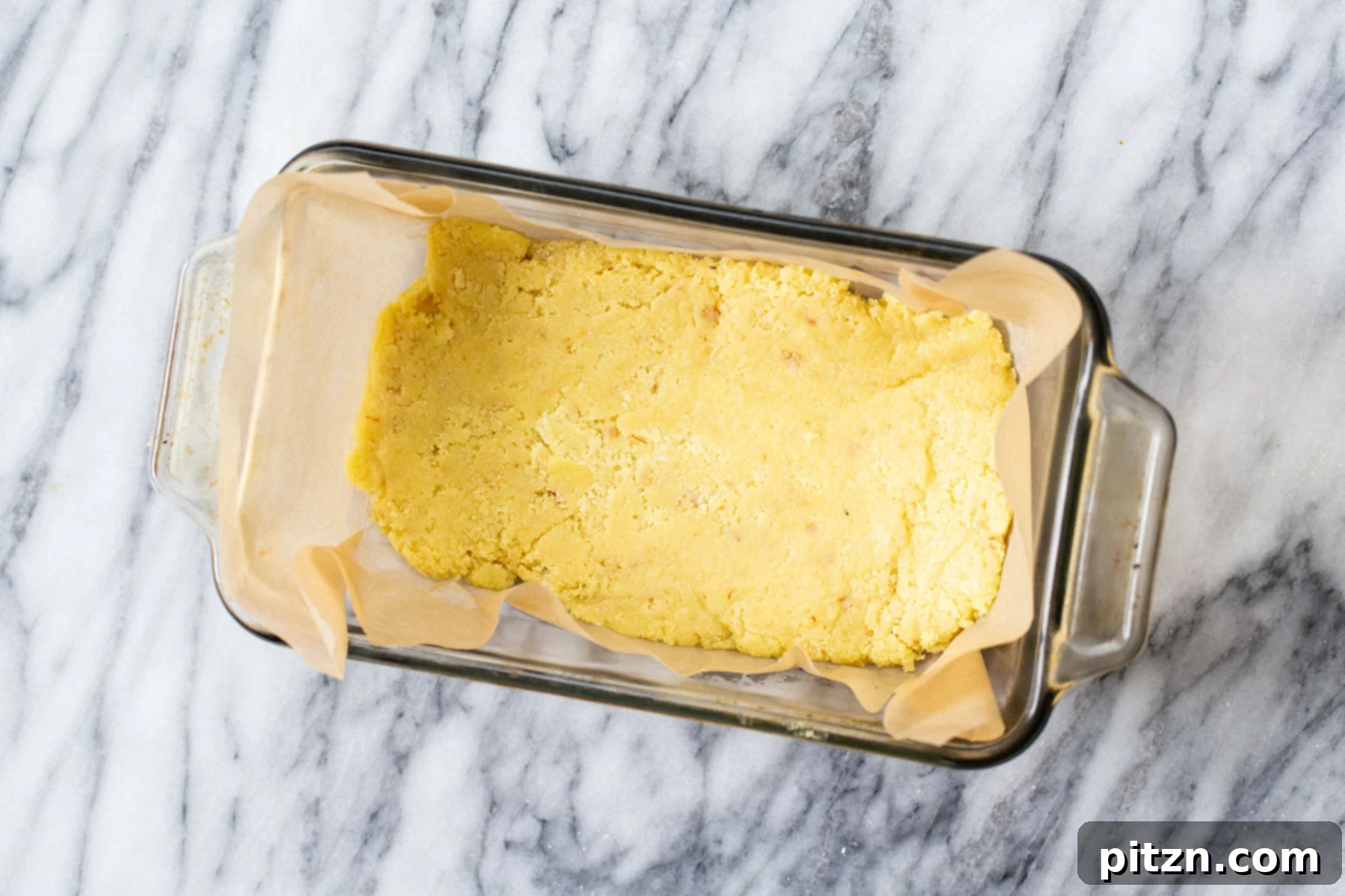 Rich Plant-Based Almond Burfi 6 The warm almond burfi mixture being pressed firmly into a parchment-lined Pyrex loaf pan using a spatula, ensuring an even layer.