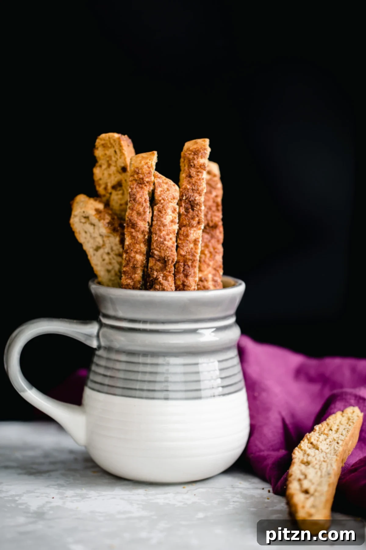 A grey and white coffee mug sits on a wooden surface, with homemade vegan churro biscotti peeking out, inviting a delightful coffee break.