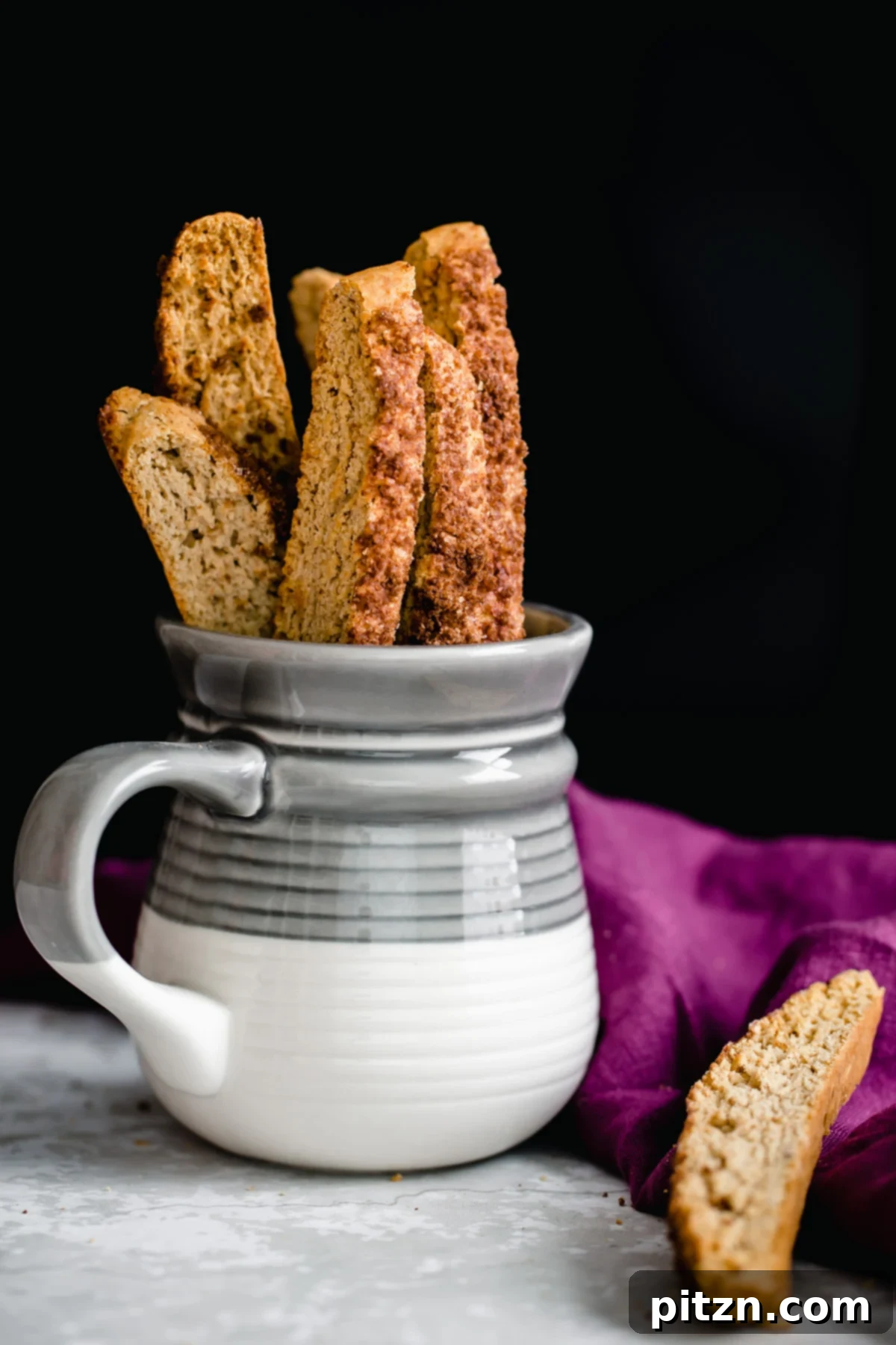 A close-up shot of homemade Vegan Cinnamon Sugar Biscotti standing upright in a small white coffee mug, highlighting their golden-brown color and cinnamon sugar coating.