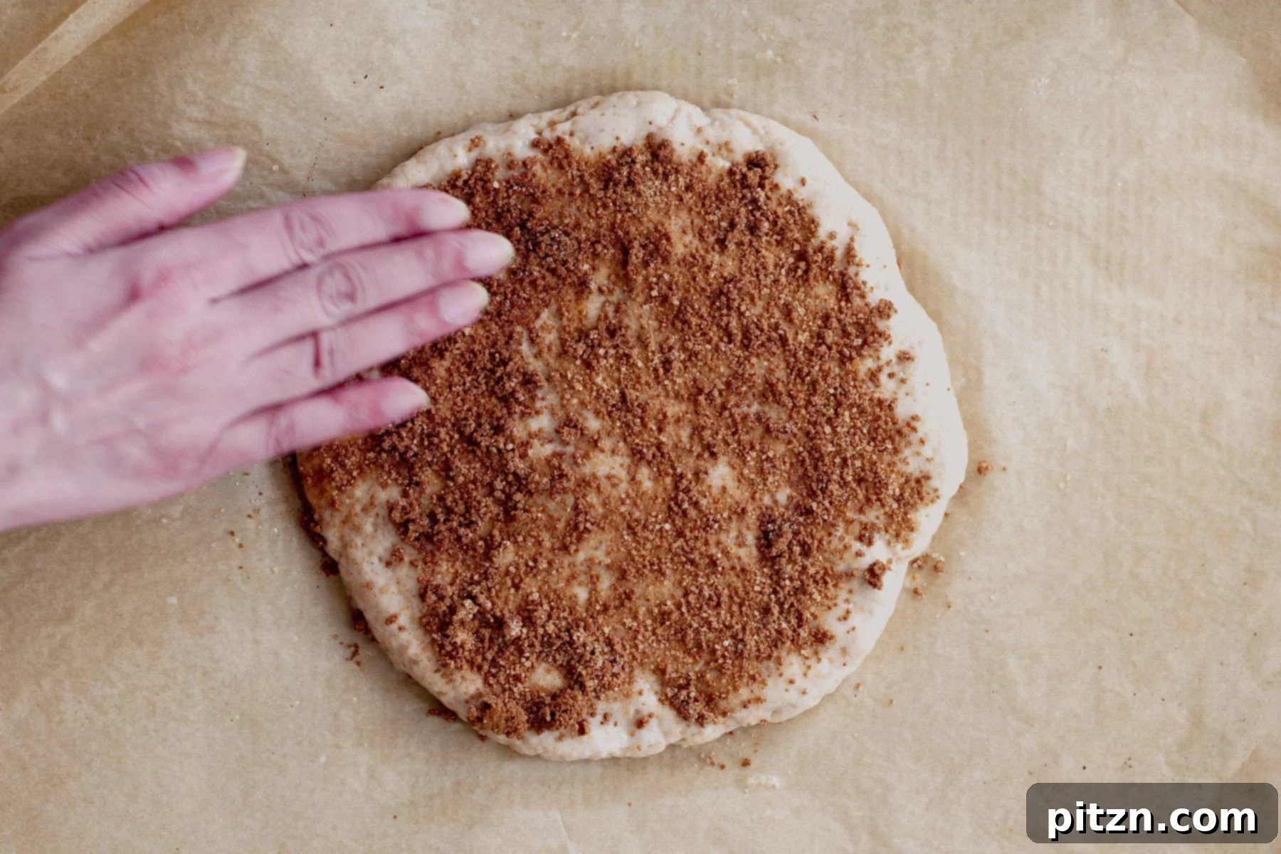 A hand sprinkles cinnamon sugar generously over a shaped loaf of biscotti dough on parchment paper, preparing it for baking.