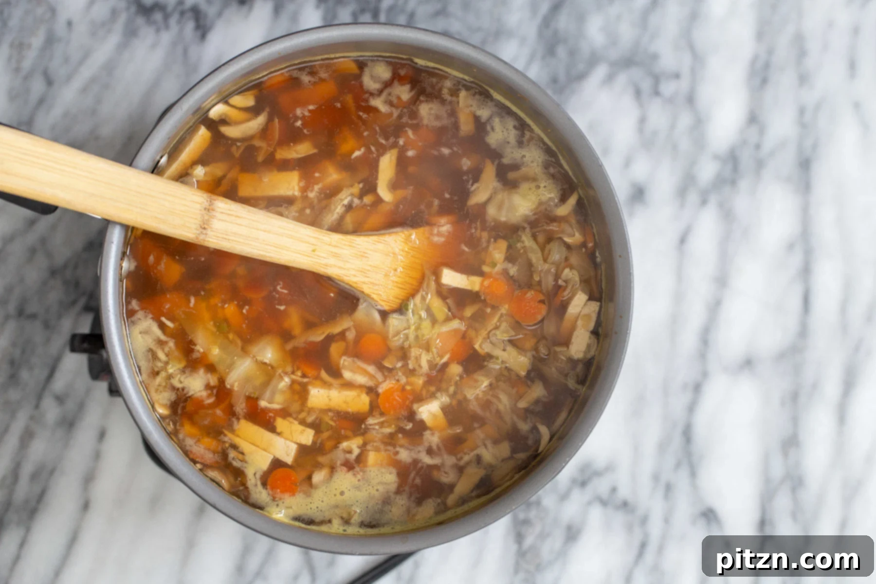 Vegan Hot and Sour Ramen Bowl 9 A wooden spoon stirs the rich, reddish broth of vegan hot and sour soup, blending the vegetables and aromatics.