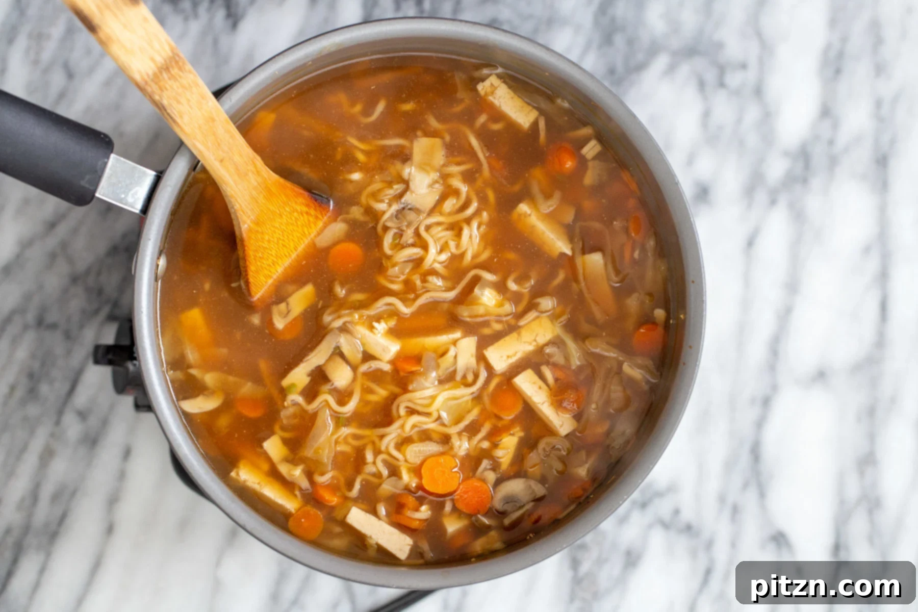 Vegan Hot and Sour Ramen Bowl 13 A silver soup pot filled with delicious Vegan Hot and Sour Ramen Soup, with a wooden spoon resting inside, ready for serving.