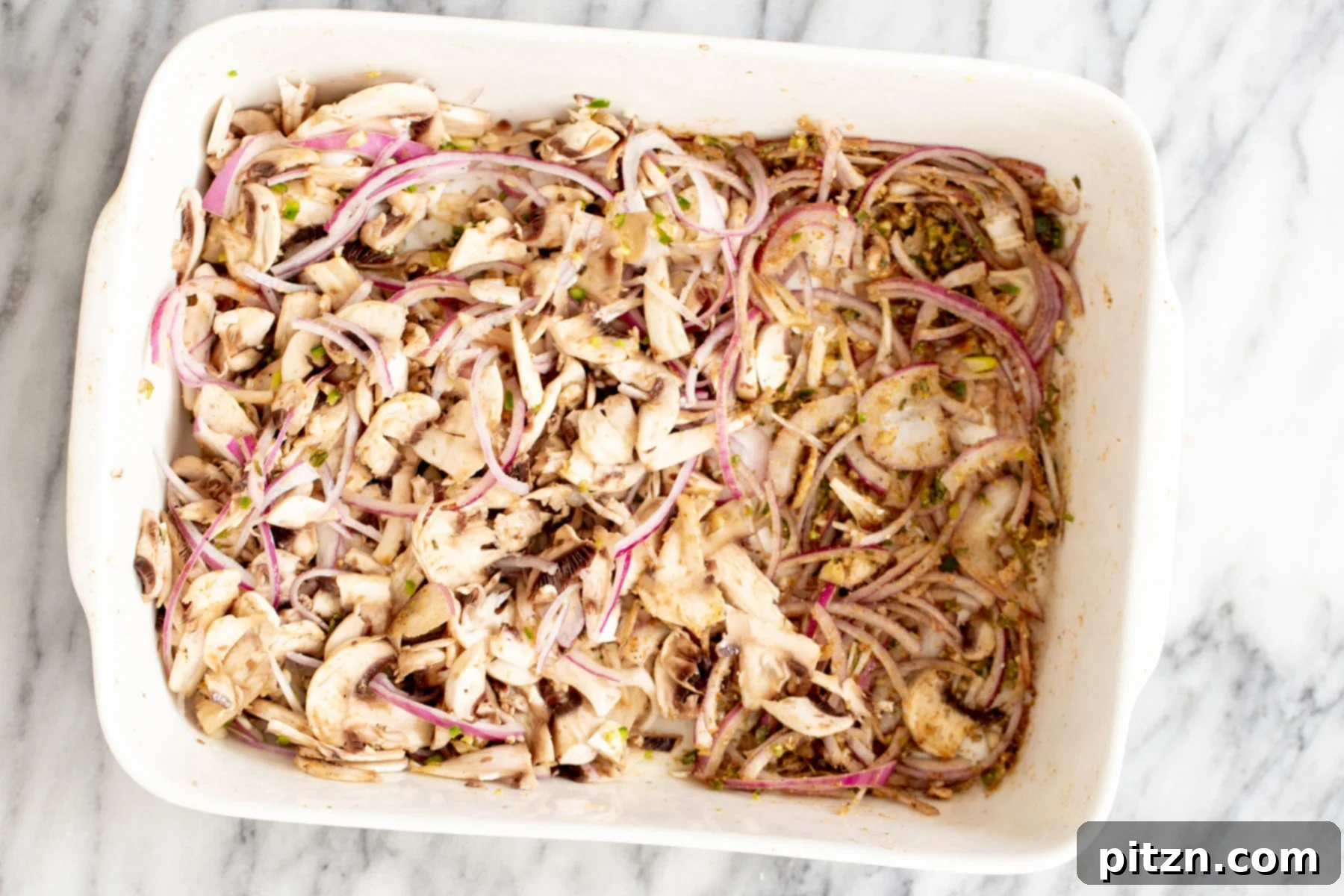 Sliced onions, mushrooms, chili, and garlic spread out in a casserole dish before baking.