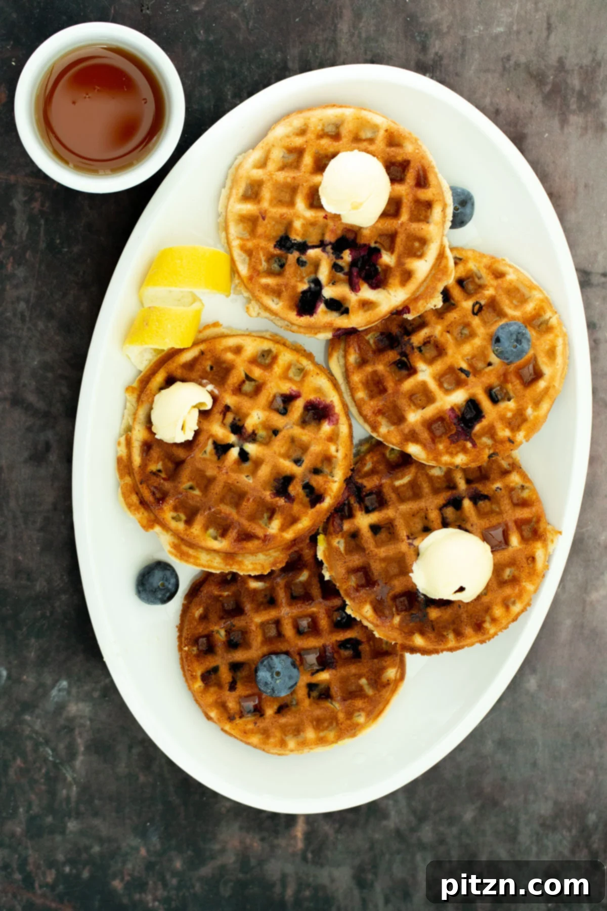 A pristine white platter elegantly displays several vegan gluten-free lemon blueberry waffles, artfully arranged and served with a small pitcher of rich maple syrup on the side, inviting a delightful breakfast.