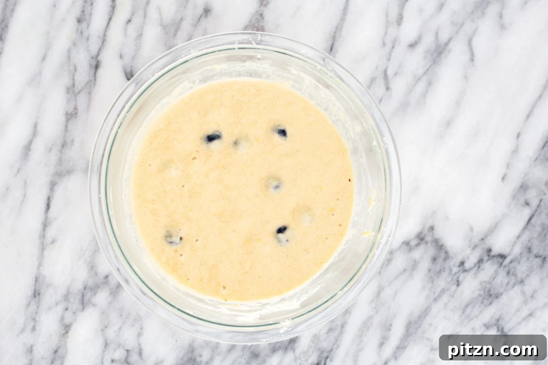 A clear glass bowl filled with a smooth, thick gluten-free waffle batter, speckled with fresh blueberries and delicate lemon zest, waiting to be cooked.