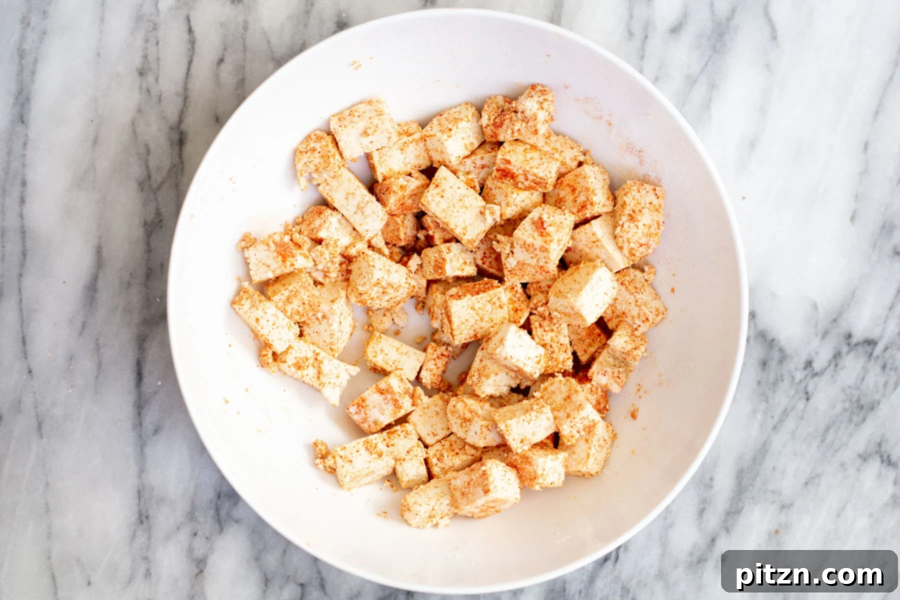 Vegan Tofu Rogan Josh 9 Tofu cubes being tossed with a cornstarch and spice mix in a white bowl, ensuring even coating.