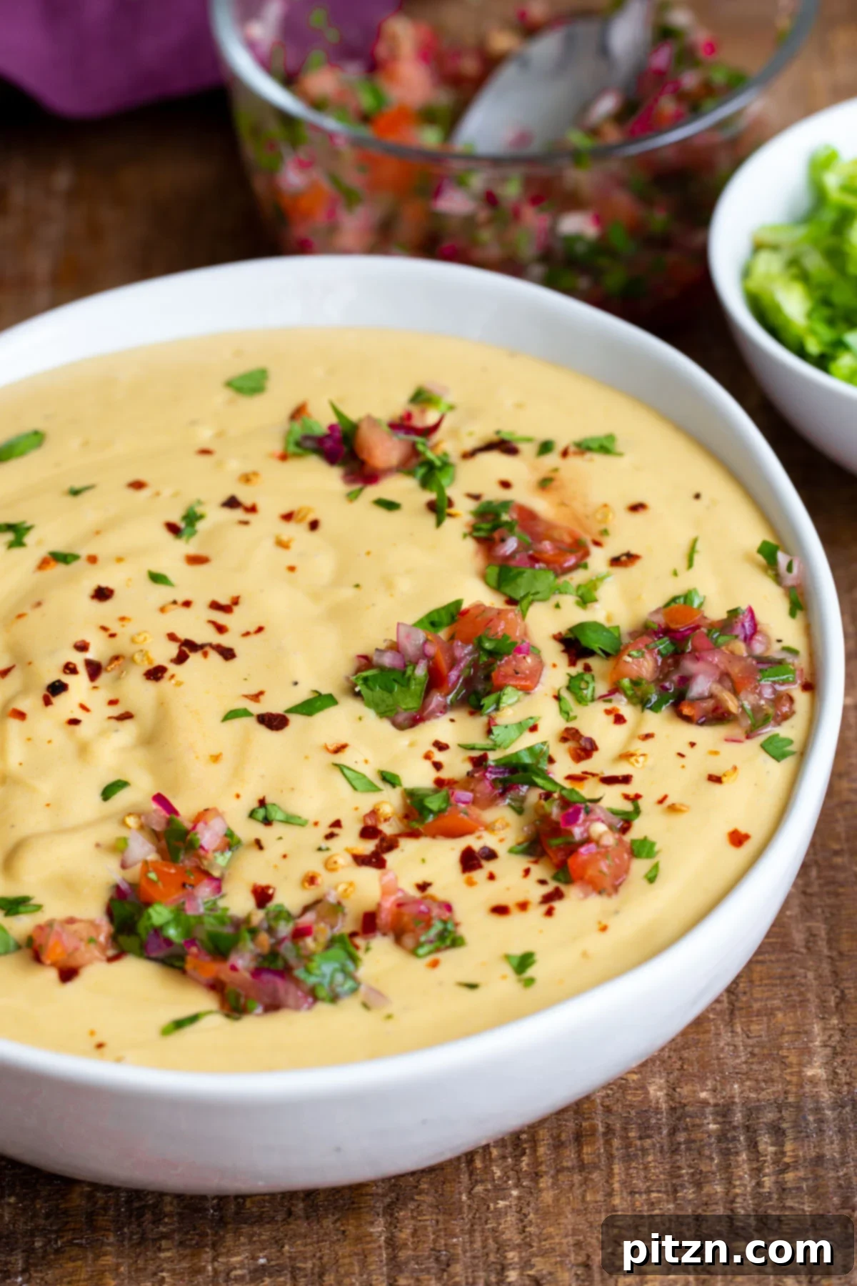 Side view of a white bowl filled with creamy vegan cheese sauce, generously sprinkled with fresh herbs and chopped tomatoes.