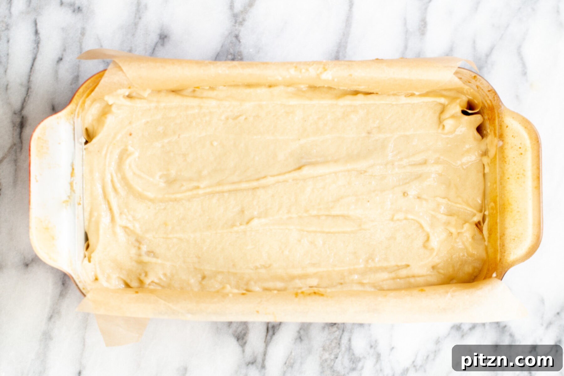 A loaf pan filled with layers of cake batter and baklava topping, sitting on a pristine marble counter.
