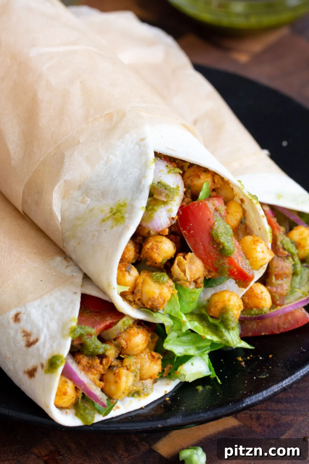 Tangy Chickpea Wraps 5 Three fully assembled chickpea kathi rolls, wrapped in parchment paper, served on a dark background. One is unwrapped to show the vibrant chickpea filling, tomatoes, and onions.