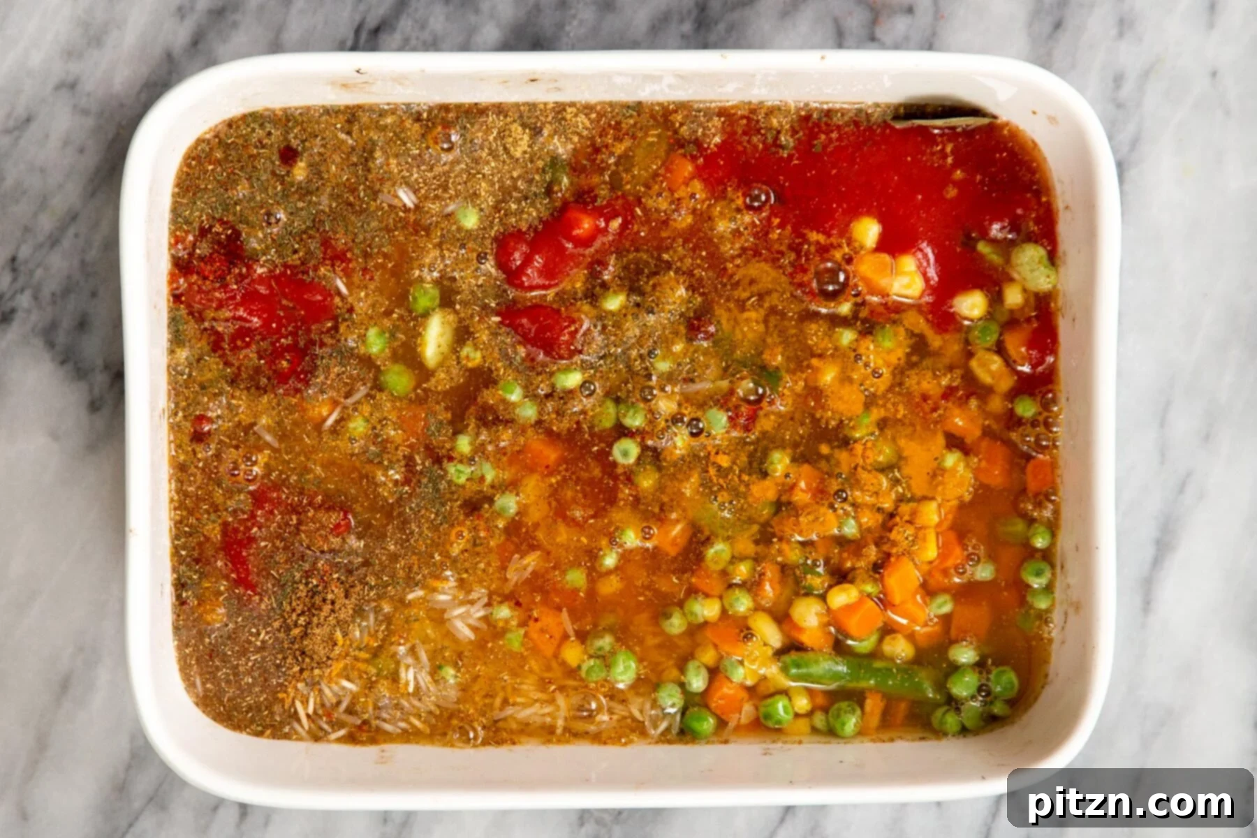 All dry and liquid ingredients for the baked masala rice pulao combined in a white casserole dish, awaiting baking.