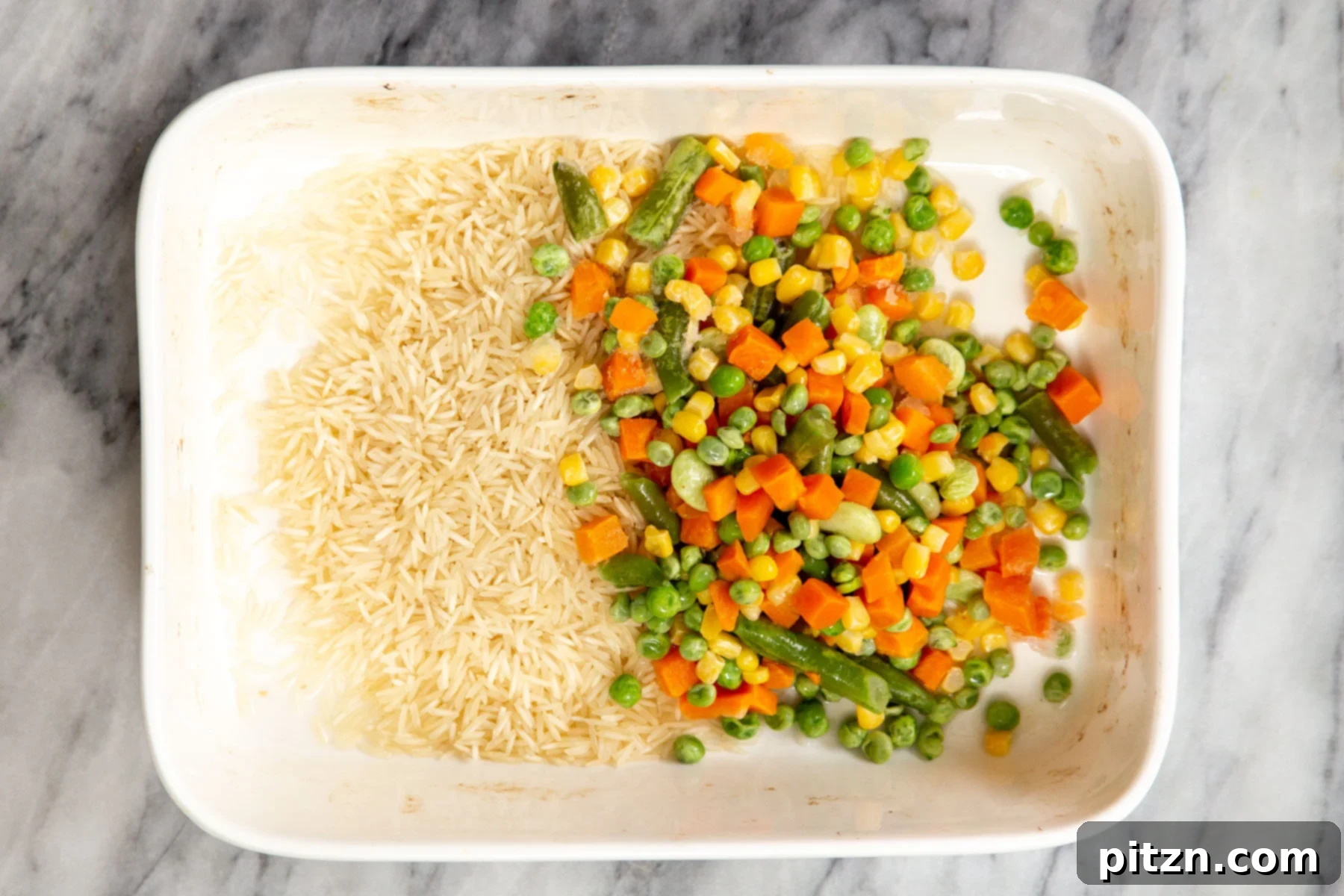 Uncooked basmati rice and frozen mixed vegetables placed in a white casserole dish, ready for seasoning.