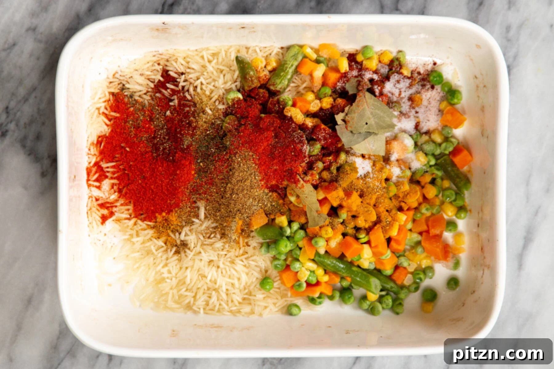 A white casserole dish with uncooked rice, a medley of colorful spices, and mixed vegetables, ready to be combined.