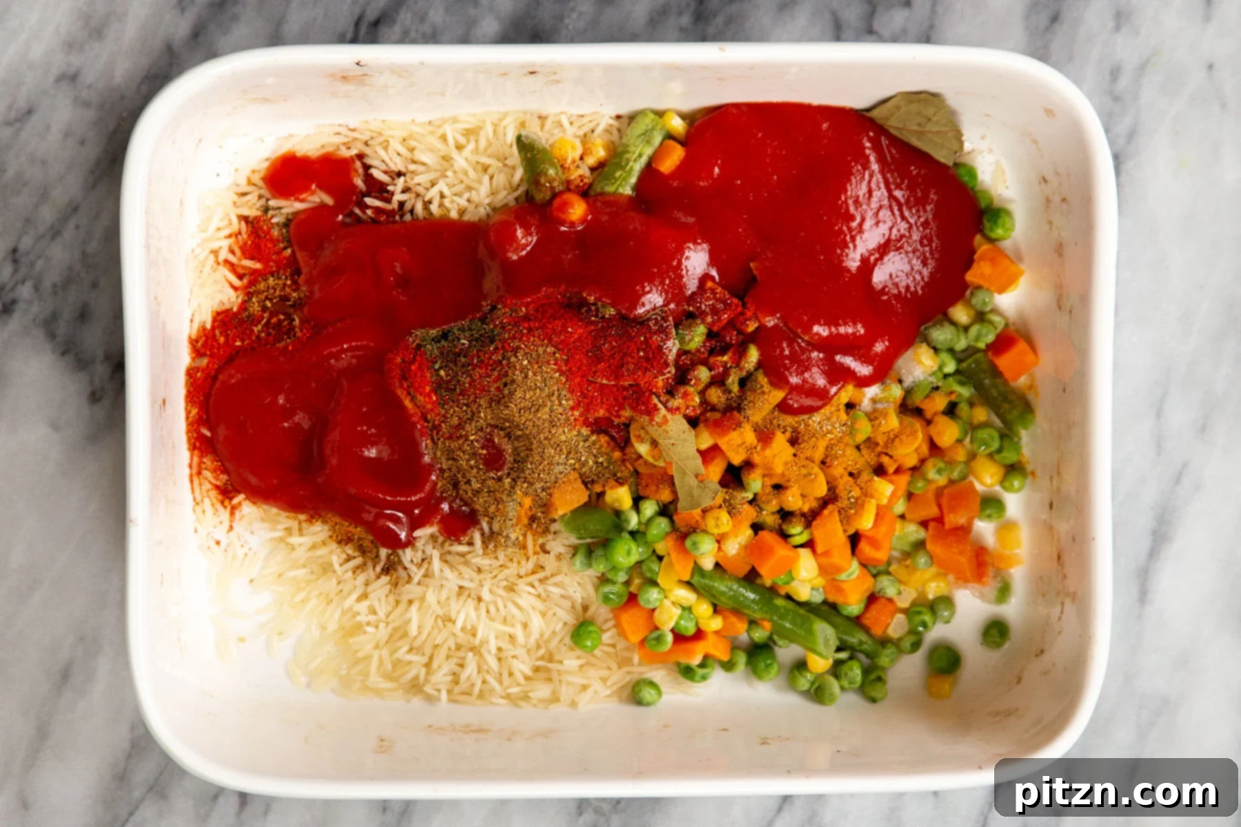 A white casserole dish containing rice, rich tomato paste, fresh vegetables, and an array of fragrant spices, all mixed together.