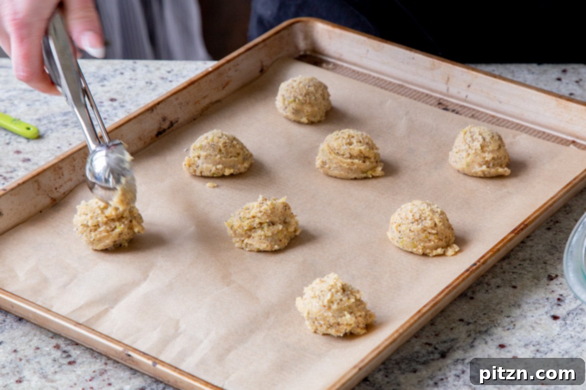 A hand scooping balls of vegan lemon poppyseed cookie dough onto a parchment-lined baking sheet, meticulously spacing them out before baking.