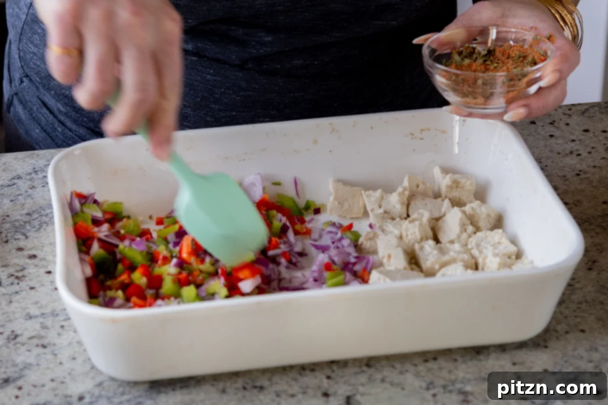 Fragrant Tikka Masala Rice Bake with Golden Crispy Tofu 12 A hand stirring a vibrant spice blend into chopped vegetables (onions, bell peppers, green chili) in a baking pan.