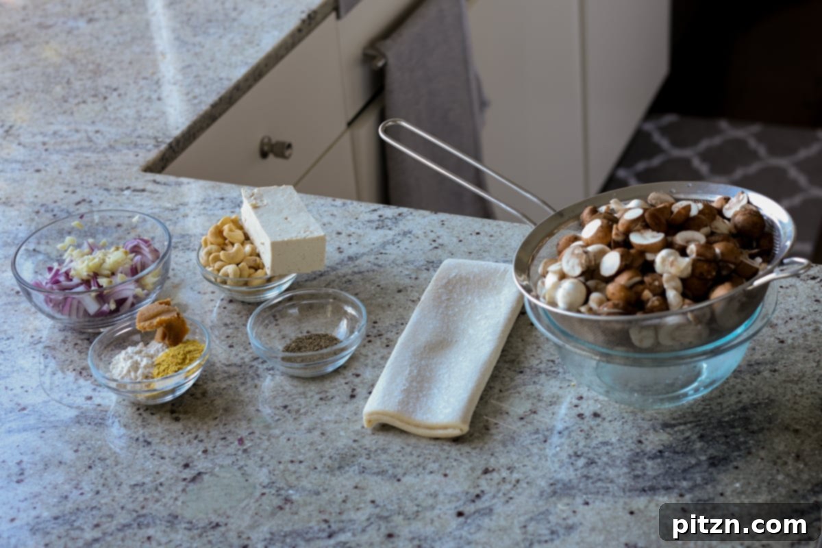 Assortment of fresh ingredients for mushroom puffs laid out on a kitchen counter: mushrooms, onions, garlic, cashews, tofu, herbs, and puff pastry.