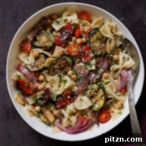A serving bowl of refreshing Summer Pasta Salad with grilled vegetables and a serving spoon. The salad is vibrant and colorful.